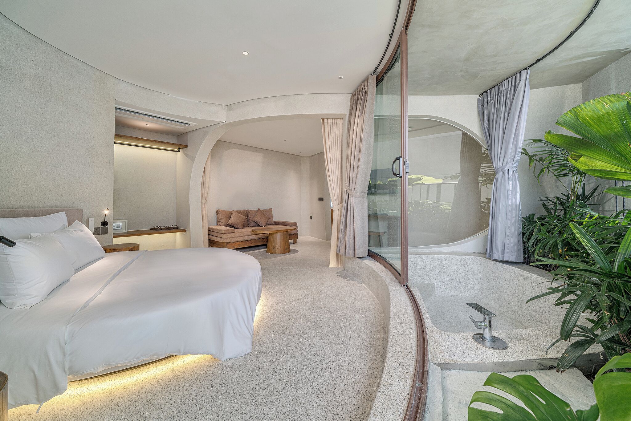 A modern hotel room with a large bed, a sitting area with a sofa and coffee table, a glass-enclosed jacuzzi, and a large potted plant.