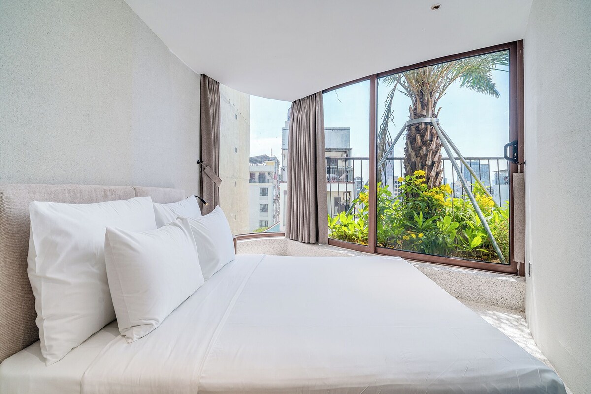 A hotel room with a large bed, a balcony with a view of buildings and greenery.