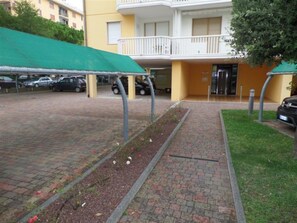 Ein gepflasterter Weg unter einem grünen Blätterdach, eine Rasenfläche und ein Gebäude mit Balkon im Hintergrund.