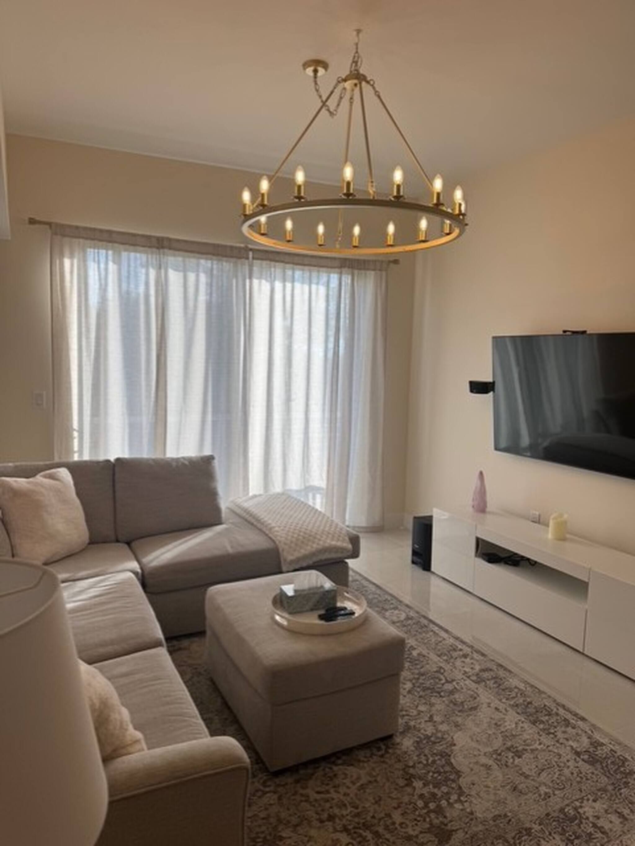 A modern living room with a sectional sofa, a flat-screen TV, a chandelier, and a large window with curtains.