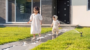 Dos niños jugando con agua en un jardín frente a un edificio moderno.