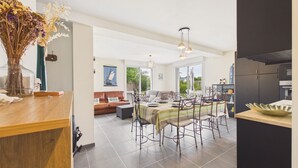 Un'ampia sala da pranzo con un lungo tavolo e sedie, una cucina con pensili scuri e una zona living con divano e un quadro.
