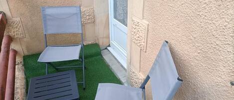 Dos sillas plegables y una mesita sobre césped artificial, con una pared beige texturizada y una ventana con vidrio esmerilado.