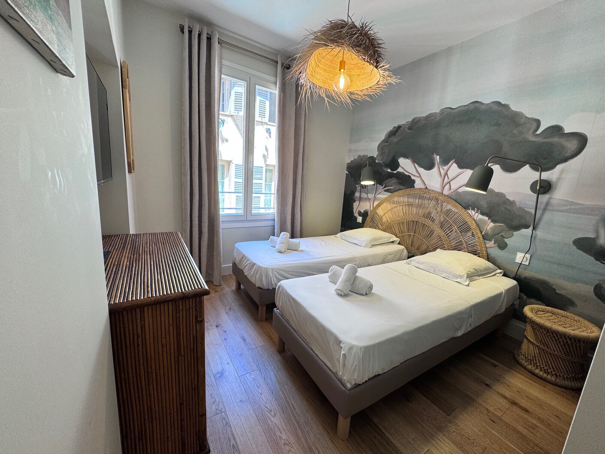 A bedroom with two beds, a wooden bench, a wicker chair, a wall-mounted lamp, and a window with curtains.