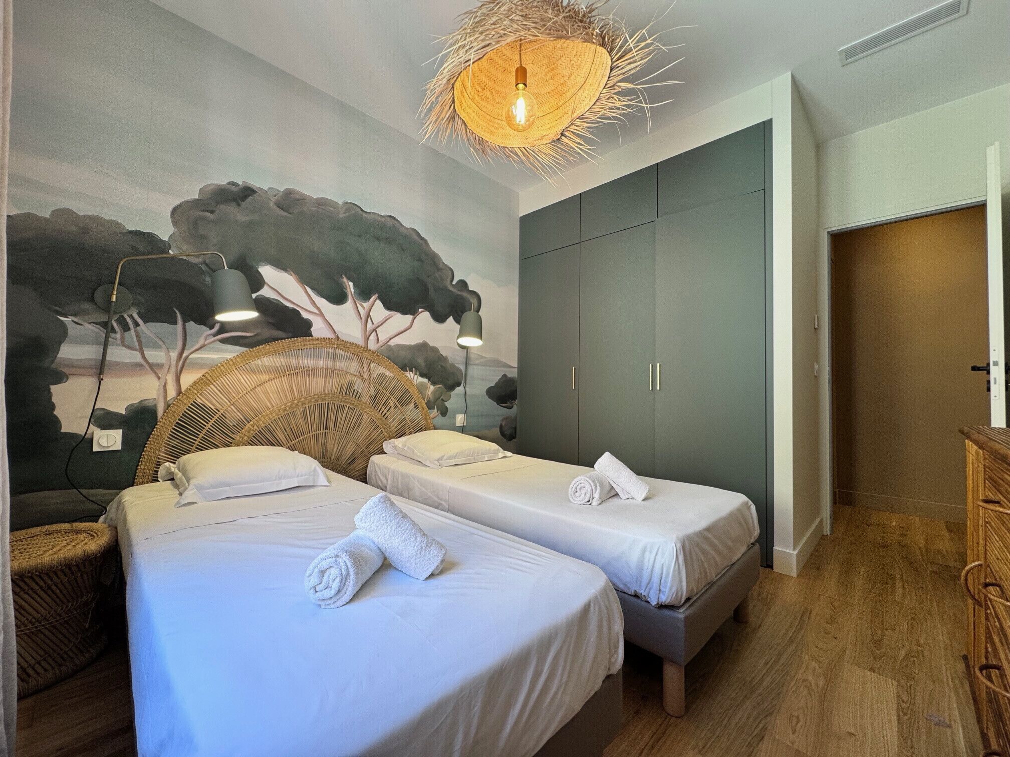 Ein Zimmer mit zwei Betten, Wandgemälde, Korbsessel und Kleiderschrank.