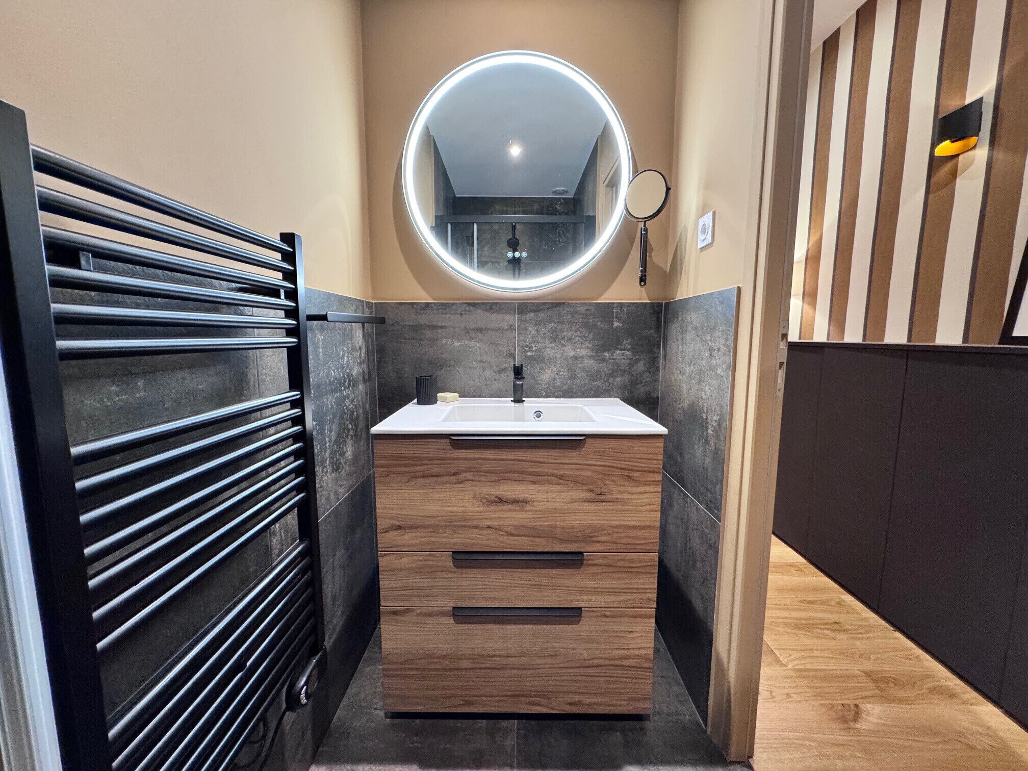Une salle de bain moderne avec un meuble-lavabo en bois, un miroir rond avec un éclairage intégré et un porte-serviettes.