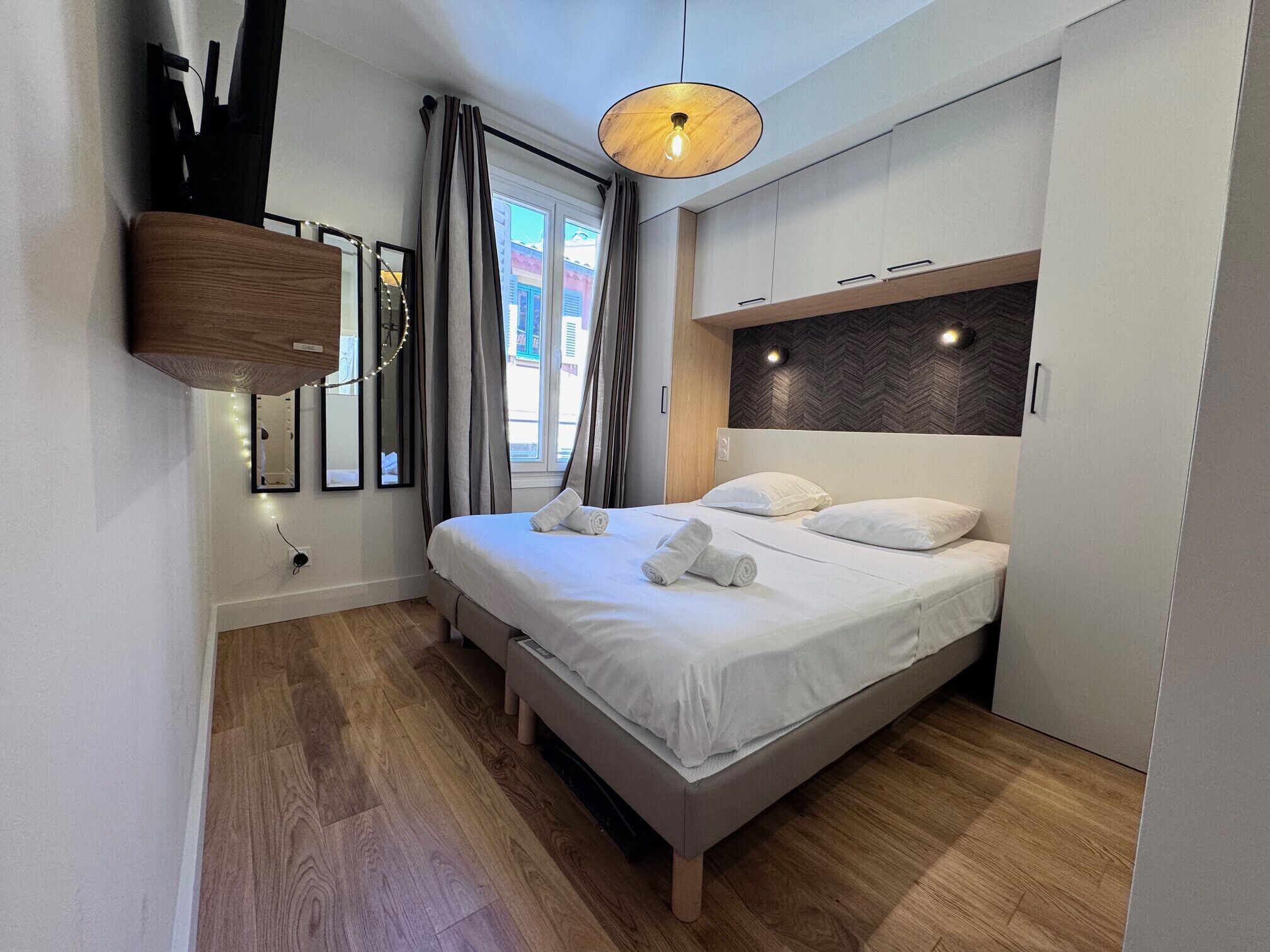 Ett modernt hotellrum med en säng, trägolv, en sänggavel med inbyggd belysning och ett fönster med gardiner.
