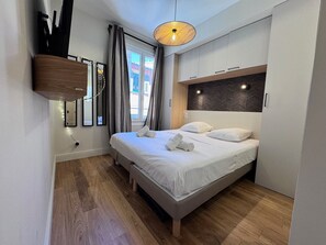 Ein modernes Hotelzimmer mit Bett, Holzboden, einem Kopfteil mit integrierter Beleuchtung und einem Fenster mit Vorhängen.
