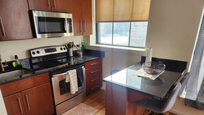 A compact kitchen with wooden cabinets, a microwave, stove, and a small dining area with a chair. - Apartment in downtown Newark, walking to Penn Sta. (Newark)
