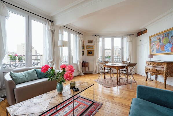 Ein Wohnzimmer mit einer Couch, einem Couchtisch und Sesseln, ein Esstisch mit Stühlen sowie große Fenster mit Vorhängen.