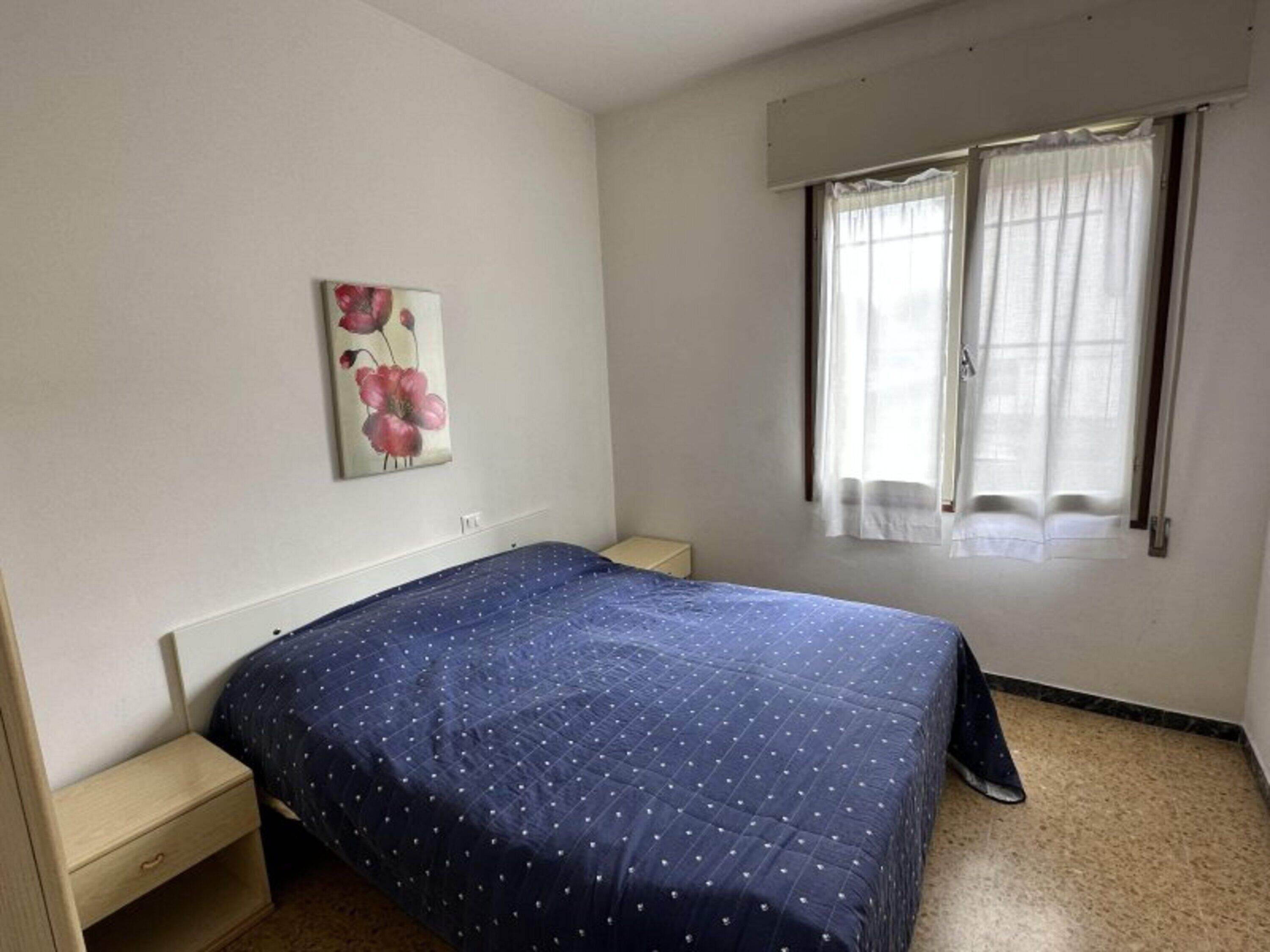 Una camera da letto con un letto, un comodino, una finestra con tende e un quadro appeso al muro.