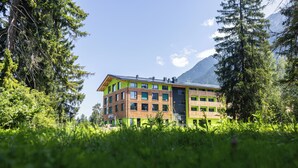 A multi-story building with a green facade, surrounded by lush greenery and mountains in the background. - Explorer Hotel Bayrischzell (Bayrischzell)