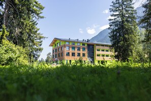 En bygning med flere etager og en grøn facade, omgivet af frodig vegetation og bjerge i baggrunden.