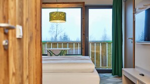 A hotel room with a bed, a desk, a television, and a balcony with a view of trees.