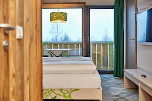 Habitación de hotel con cama, escritorio, televisión y balcón con vistas a árboles.