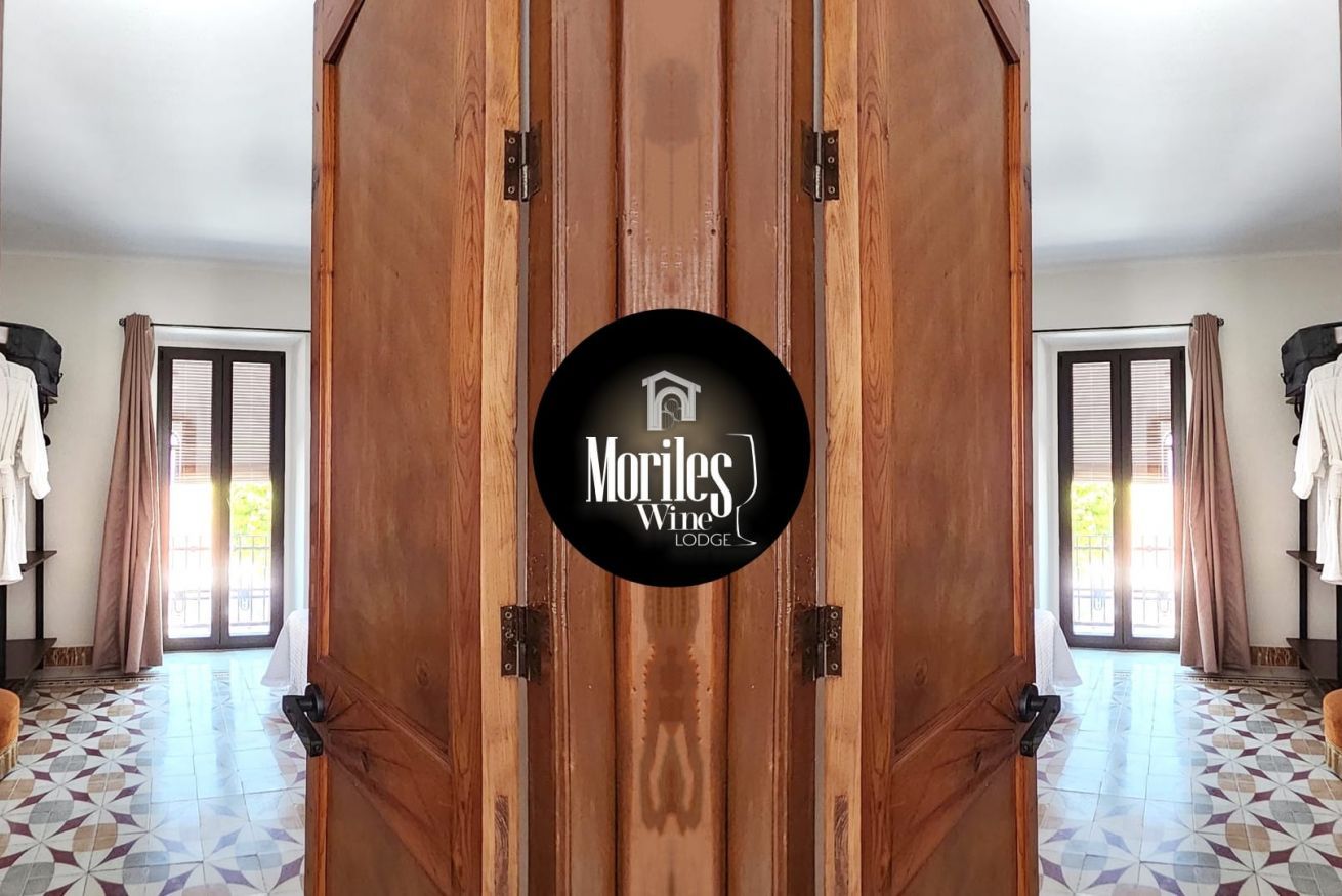 Eine Holztür mit dem Logo 'Moriles Wine Lodge' und Blick auf einen Raum mit gefliestem Boden und einem mit Vorhängen versehenen Fenster.