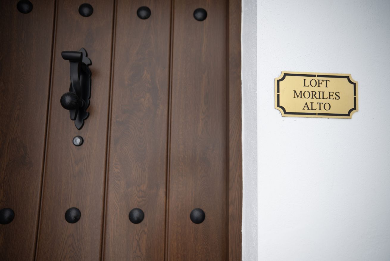 A wooden door with a black handle and a brass doorknob, and a brass plaque with the text 'LOFT MORILES ALTO' on the wall.