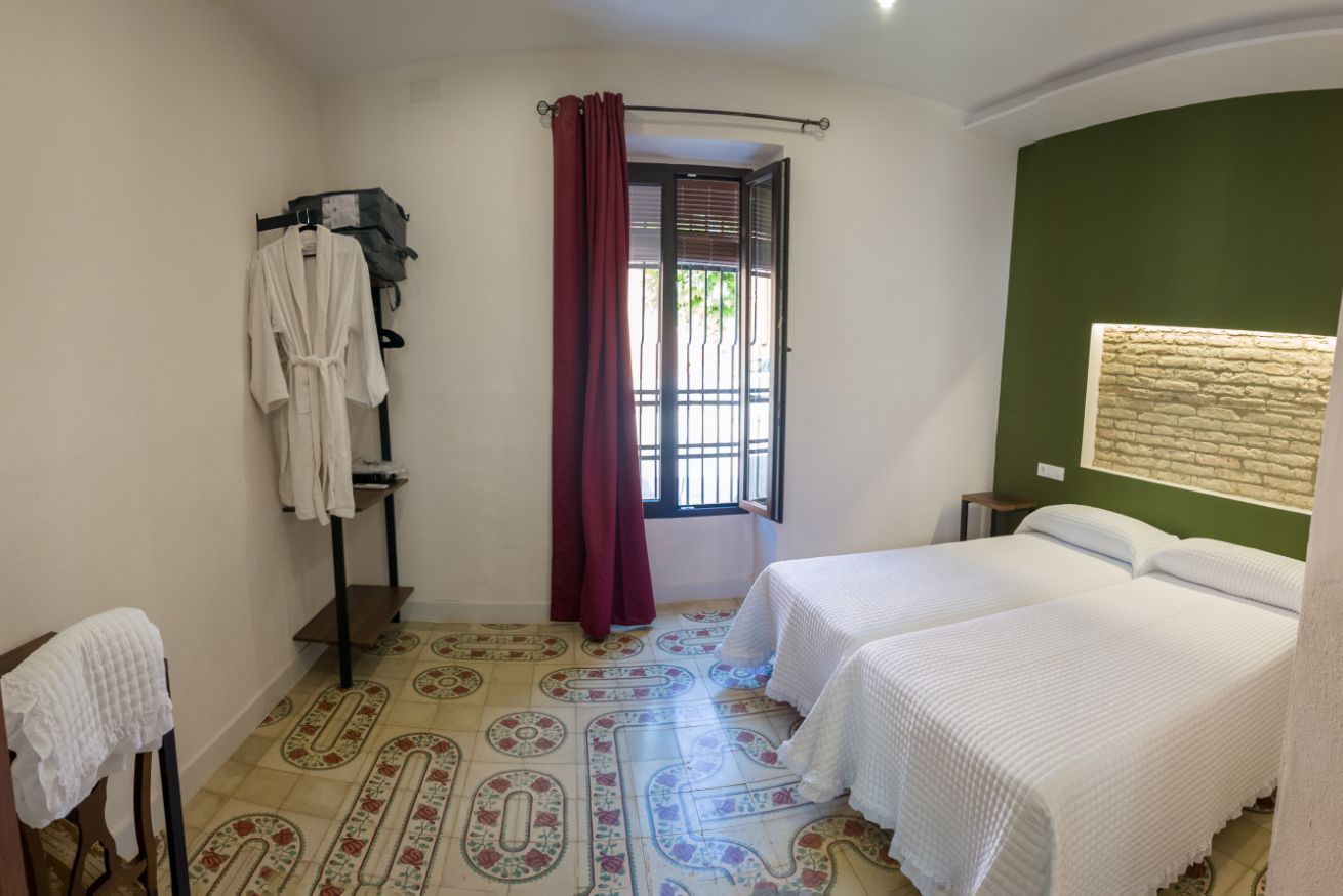 A room with two beds, a window with curtains, a towel rack, and a patterned floor.