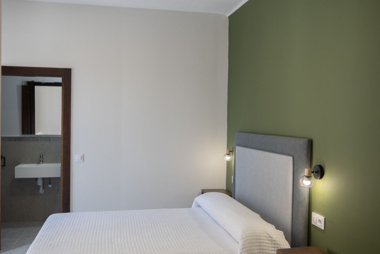 A hotel room with a bed, a nightstand, a wall-mounted lamp, and a bathroom with a sink.
