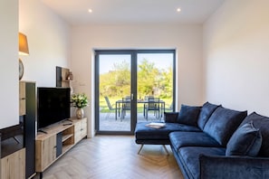 Ein modernes Wohnzimmer mit Flachbildfernseher, dunkelblauem Sofa, einer Holz-Unterhaltungseinheit und einer Glastür, die auf eine Außenpatio mit Esstisch und Stühlen führt.
