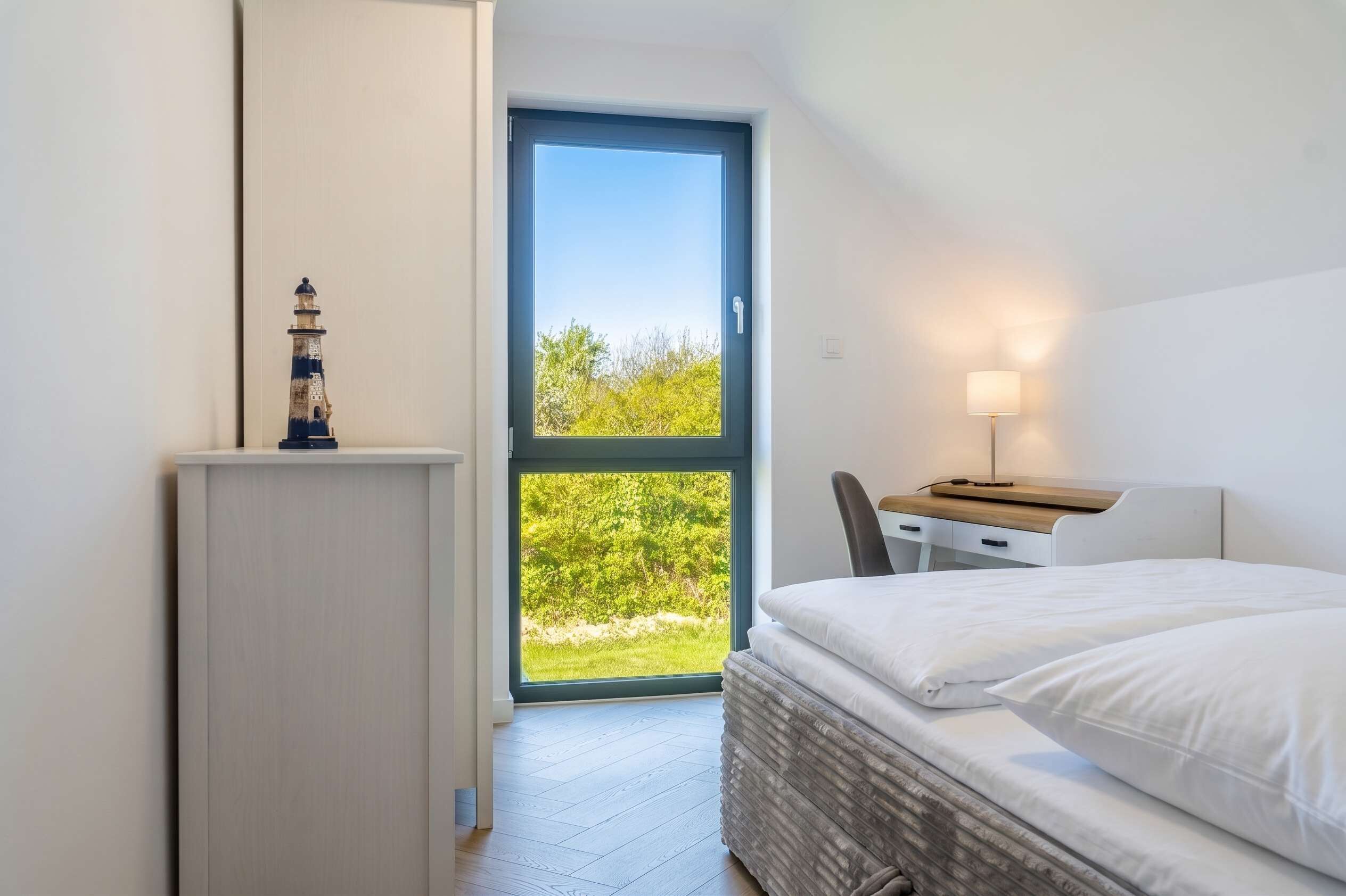 Un dormitorio con cama, escritorio, lámpara, armario y vistas a la vegetación a través de un ventanal grande.