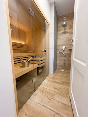 Ein zeitgemäßes Sauna-Zimmer mit Glastüren, Holzbänken, einem Metall-Eimer und einem Duschbereich mit Duschbrause.
