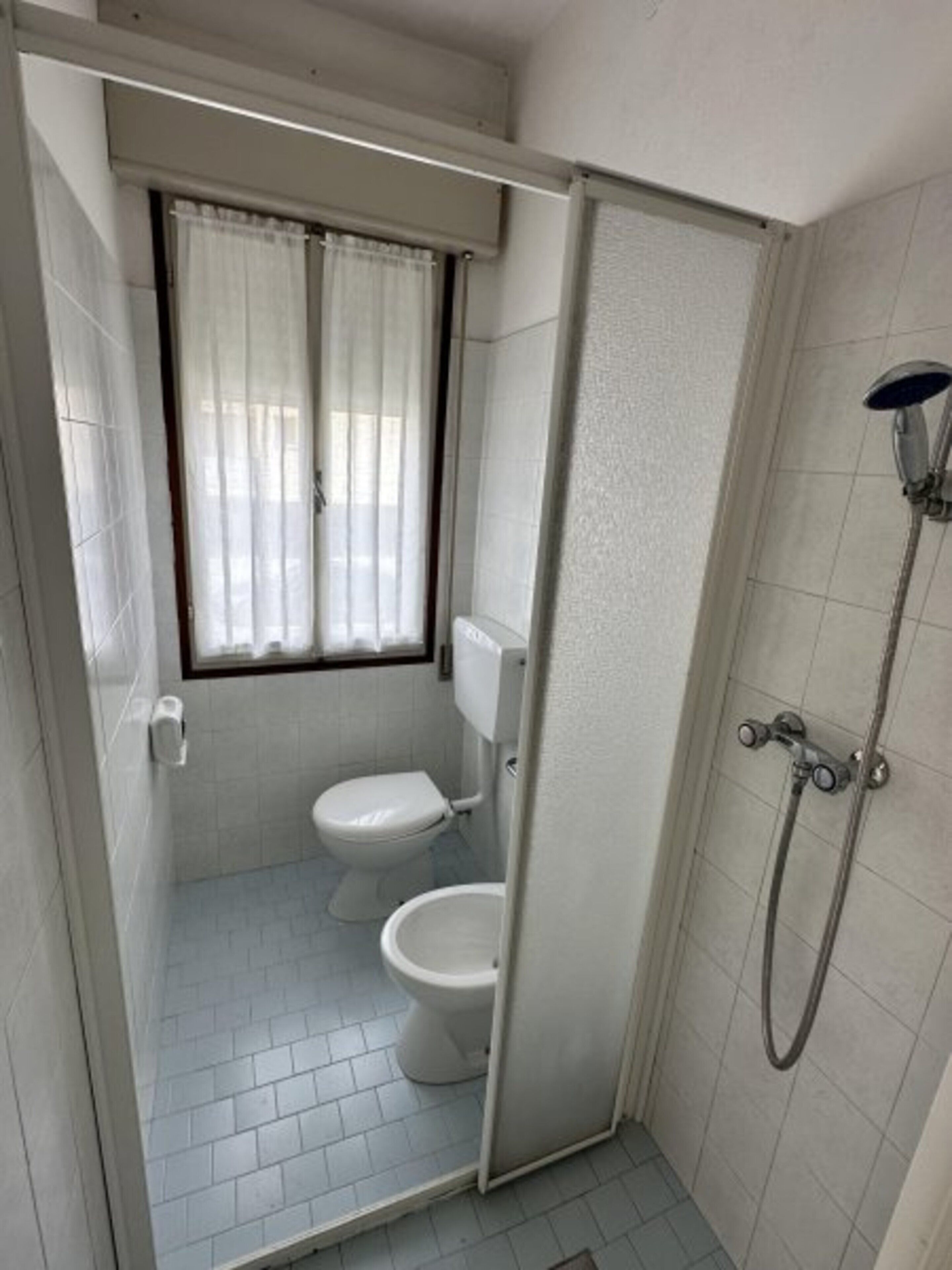 A small, enclosed bathroom with a toilet, a shower, and a window with curtains.