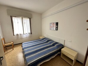 A bedroom with a bed, a chair, a bedside table, a window with curtains, and a wall-mounted picture. - Comfortable Retreat in Bibione for six Guests (San Michele al Tagliamento)