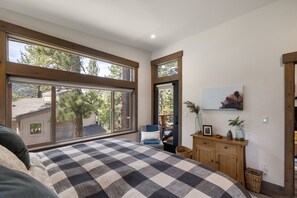 Ein Schlafzimmer mit einem großen Fenster, einem Bett mit kariertem Überwurf, einer Holzkommode und einem Bild an der Wand.
