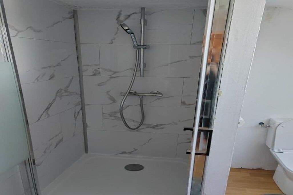 A shower with a handheld showerhead and a wall-mounted faucet, a glass door, and a tiled floor.