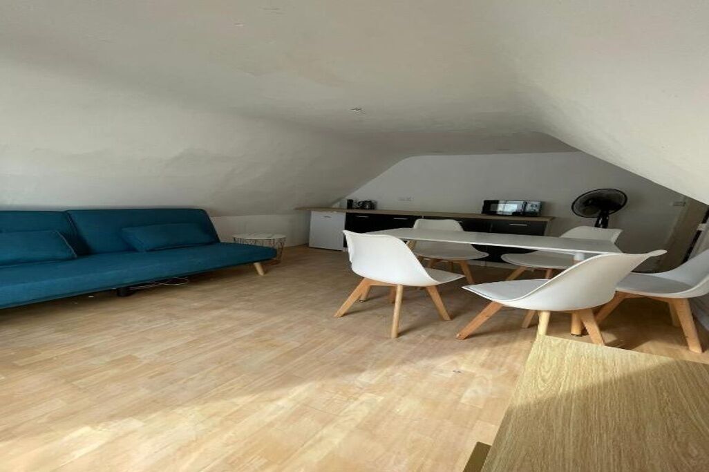 A compact living space with a blue sofa, a dining table with chairs, a kitchenette, and a fan.