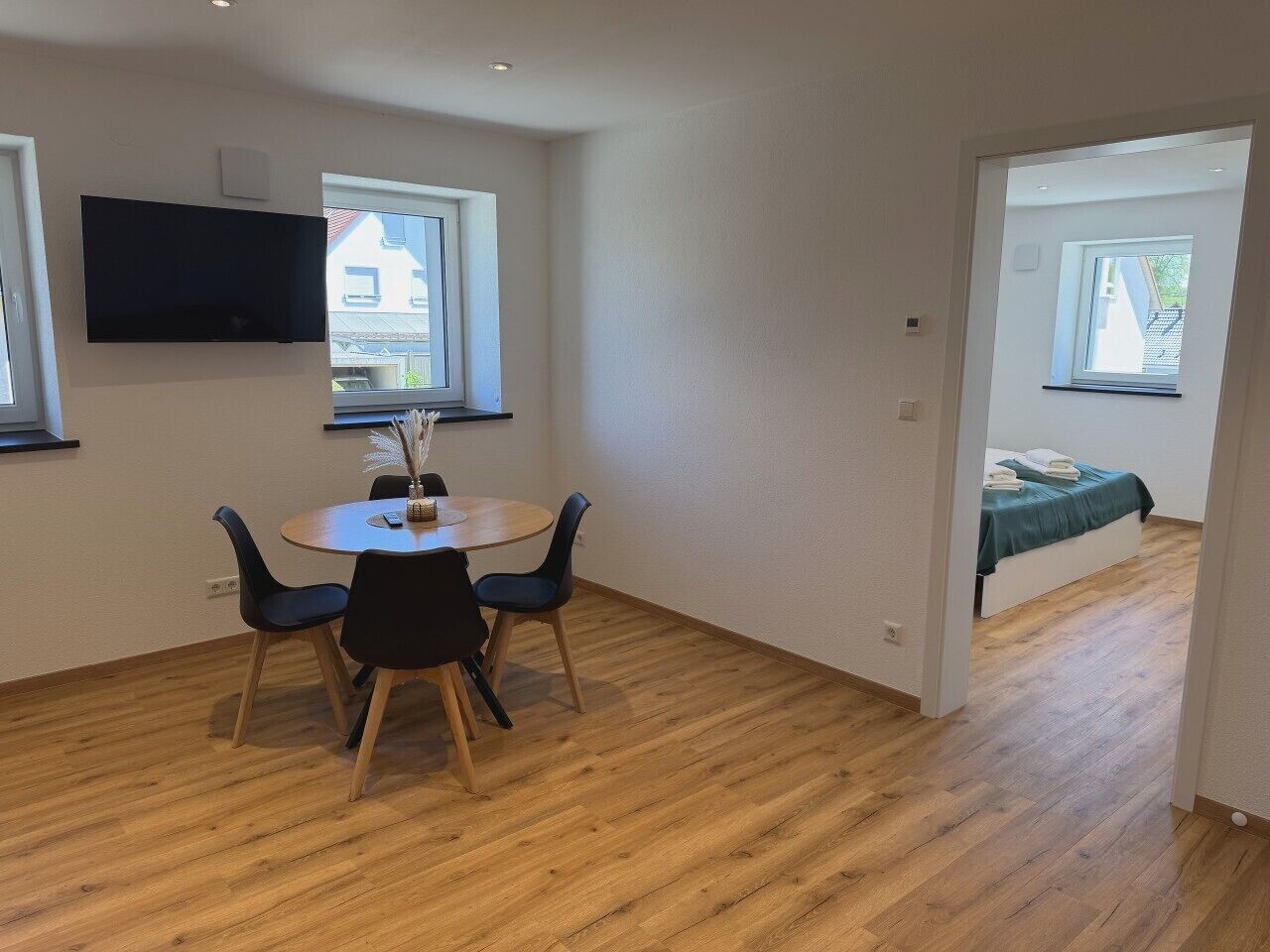 A room with a round table, chairs, a flat-screen TV mounted on the wall, and a bed with a window.