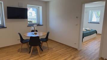 A room with a round table, chairs, a flat-screen TV mounted on the wall, and a bed with a window.
