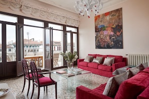 A spacious living room with a red sofa, a glass coffee table, and a view of the city through large windows. - Venice Prestige - Celestina on the Grand Canal R&R (Venice)