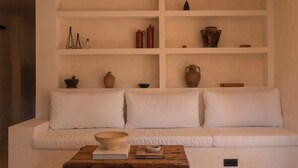 Un canapé blanc avec deux coussins, une table basse en bois sur laquelle se trouvent un bol et un livre, et un mur blanc orné d’étagères contenant divers objets.
