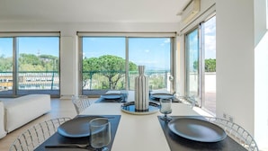 A modern dining area with a table set for four, large windows with a view of greenery and buildings, and a balcony with a railing.