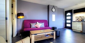 Ein modernes Wohnzimmer mit einem violetten Sofa, einem Holztisch, einer Stehlampe und einer Küchenzeile mit Schränken und einer Wanduhr.