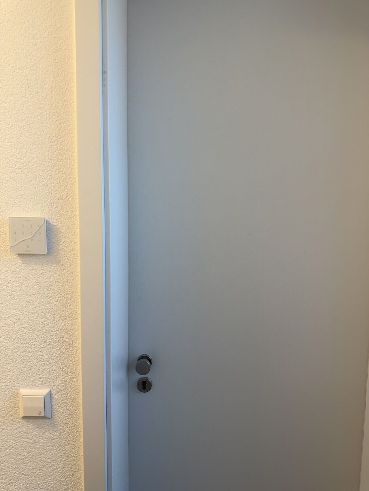 Una puerta blanca con un ojo de cerradura y un objeto pequeño y rectangular blanco a la izquierda, en la pared.
