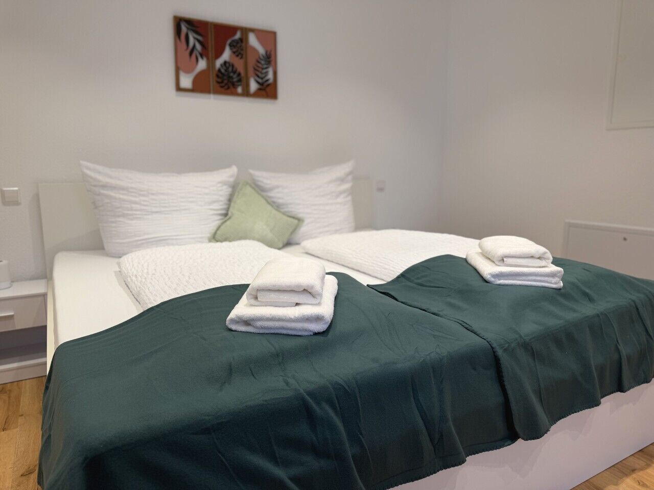 A double bed with white bedding and a green throw blanket, a nightstand with a lamp, and a wall art piece above the bed.