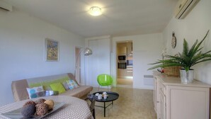 Ein Wohnzimmer mit einer Couch, einem grünen Sessel, einem kleinen Tisch mit Dekoration, einer Lampe und einer durchgehenden Tür erkennbaren Küche.
