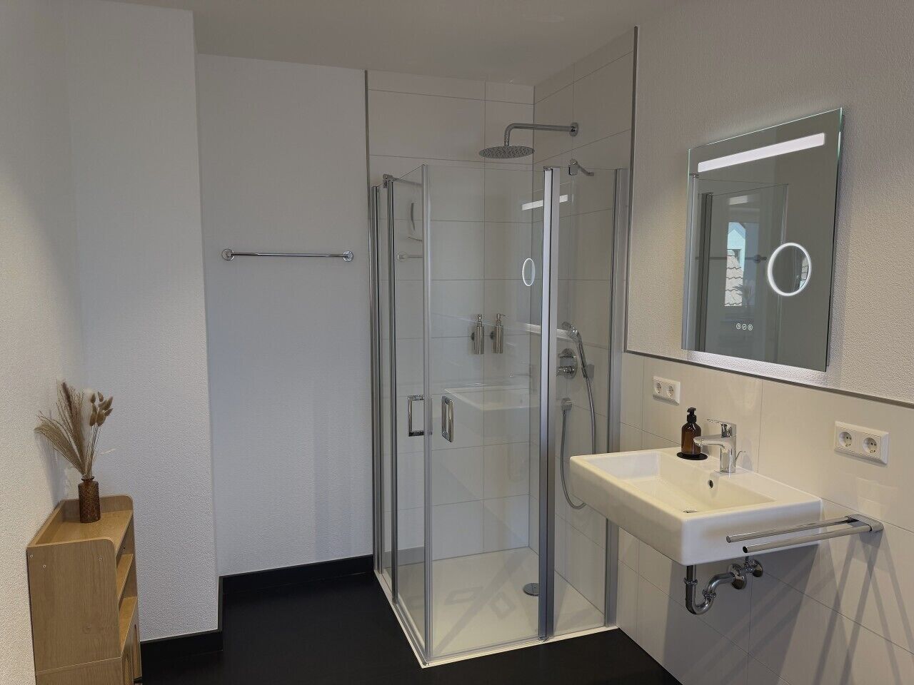 A modern bathroom with a glass-enclosed shower, a white sink with a mirror, and a wooden shelf with dried flowers.