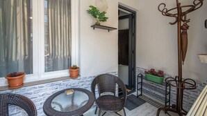 A patio area with wicker chairs, a round glass table, potted plants, a metal umbrella stand, and a door leading inside.