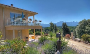 A two-story house with a balcony, surrounded by a garden with various plants and a view of mountains. - Manon - LEVIE - Gîte (LEVIE)