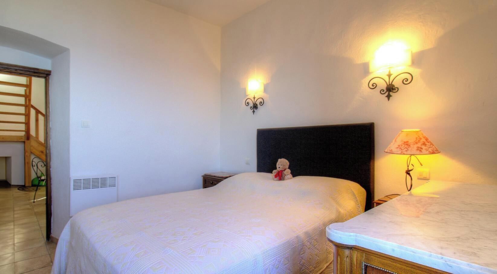 A bedroom with two beds, a headboard, bedside lamps, a teddy bear, and a staircase in the background.
