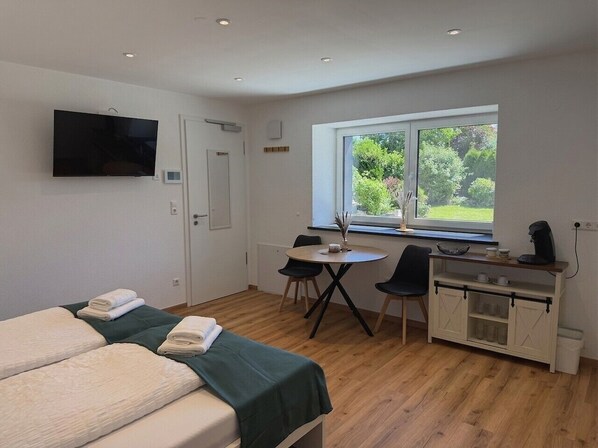 A room with two beds, a desk with chairs, a window with a view, and a kitchenette.