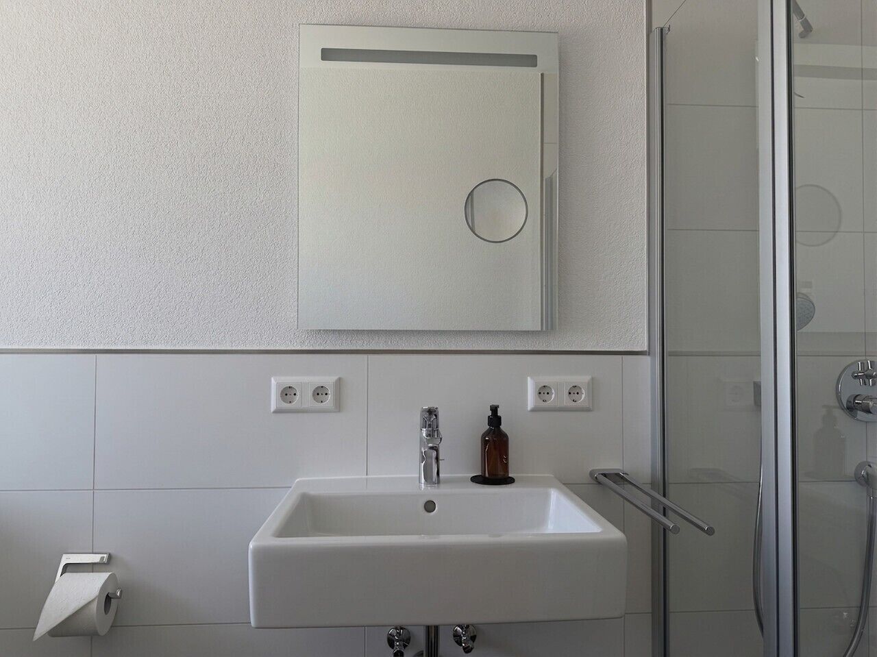 Une salle de bain avec un lavabo blanc, un miroir, une cabine de douche en verre et un distributeur de savon.