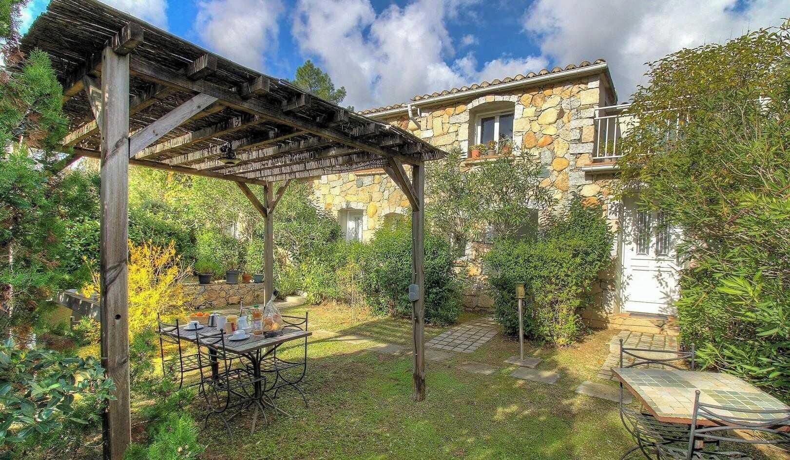 A stone building with a covered patio area, a table set for dining, and a garden with various plants.