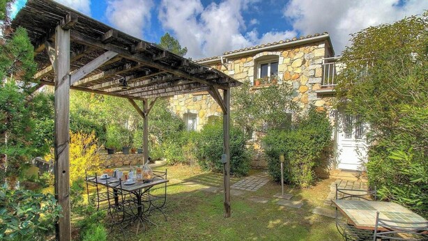 Un edificio de piedra con un patio cubierto, una mesa dispuesta para comer y un jardín con diversas plantas.