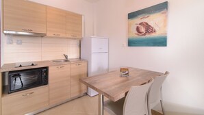 A compact kitchen with wooden cabinets, a white refrigerator, a built-in oven, and a dining area with a wooden table and two chairs.