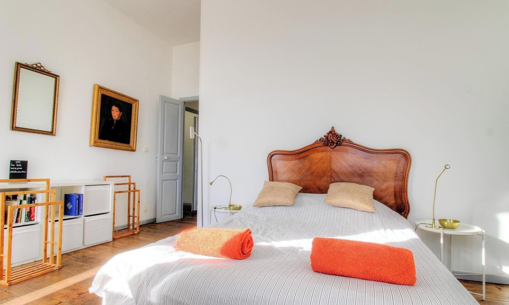 Une chambre à coucher avec une tête de lit en bois, un lit avec une literie à rayures, deux oreillers, une table de chevet avec une lampe et un bureau avec des livres et un miroir.